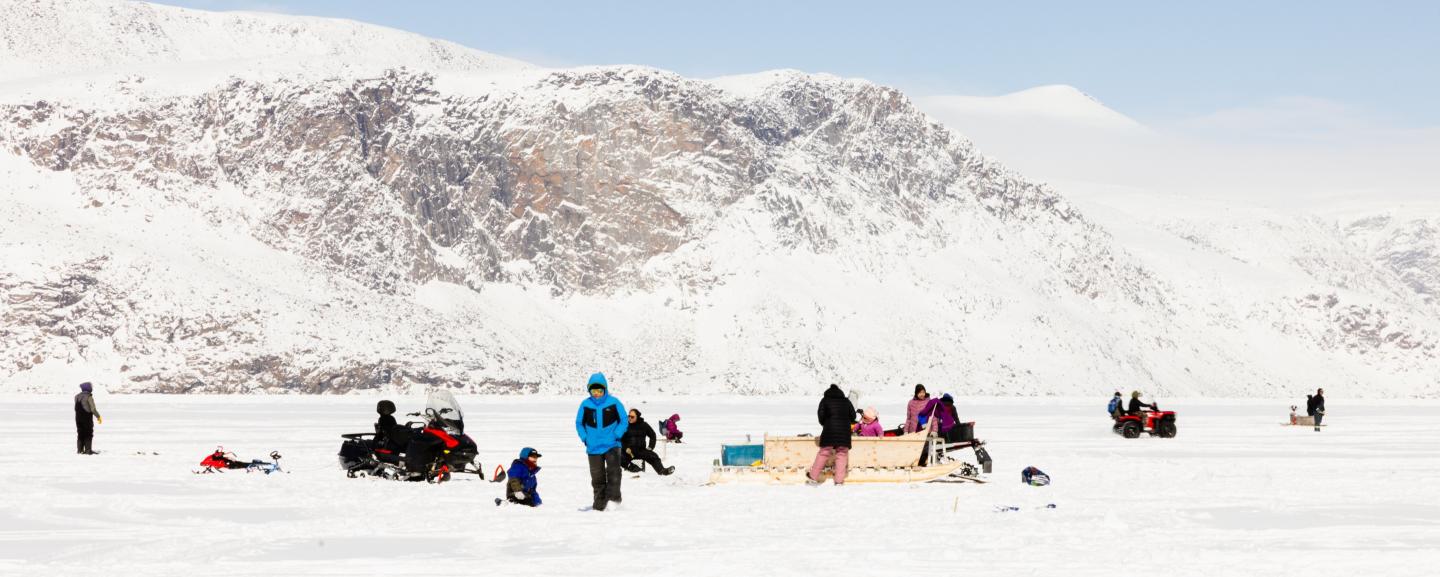 People gathered on the sea ice with snowmobiles and a qamutiik, below snow-covered cliffs under a clear blue sky.