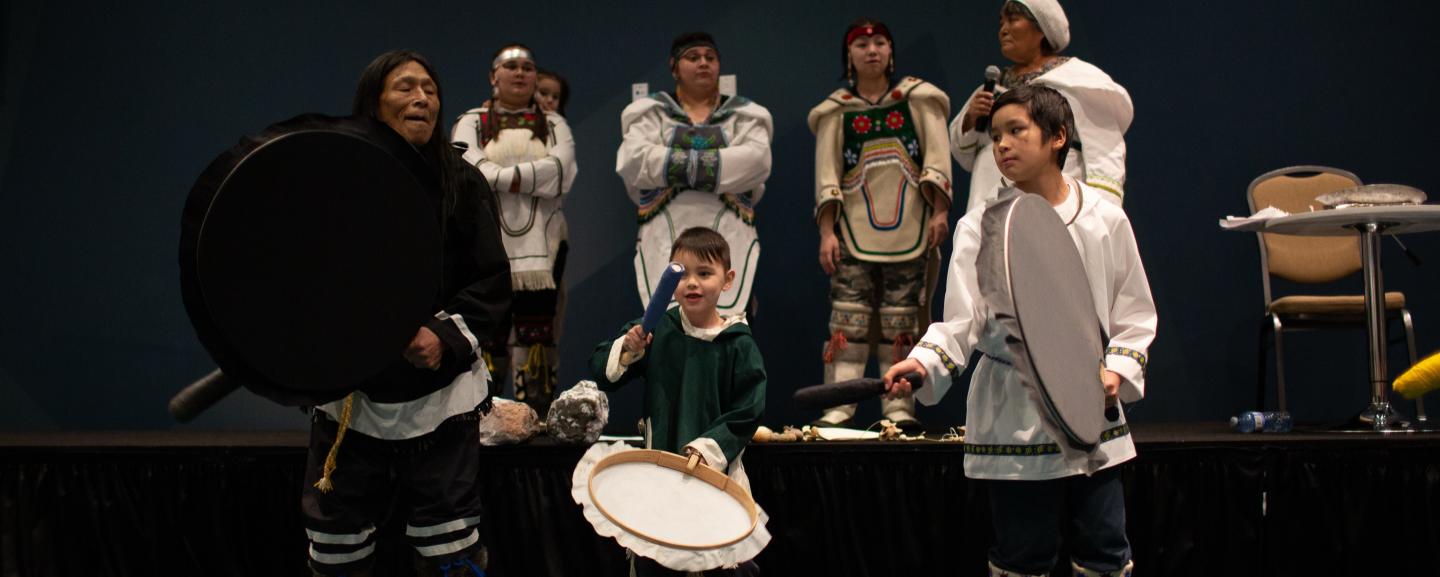 An Elder and two children drum dancing on stage, with four women in traditional clothing standing behind them.
