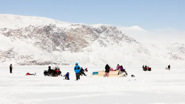 People gathered on the sea ice with snowmobiles and a qamutiik, below snow-covered cliffs under a clear blue sky.