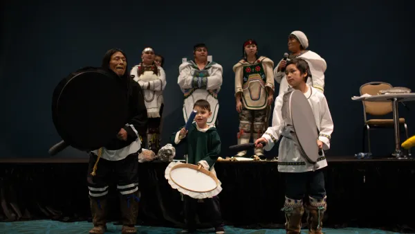 An Elder and two children drum dancing on stage, with four women in traditional clothing standing behind them.